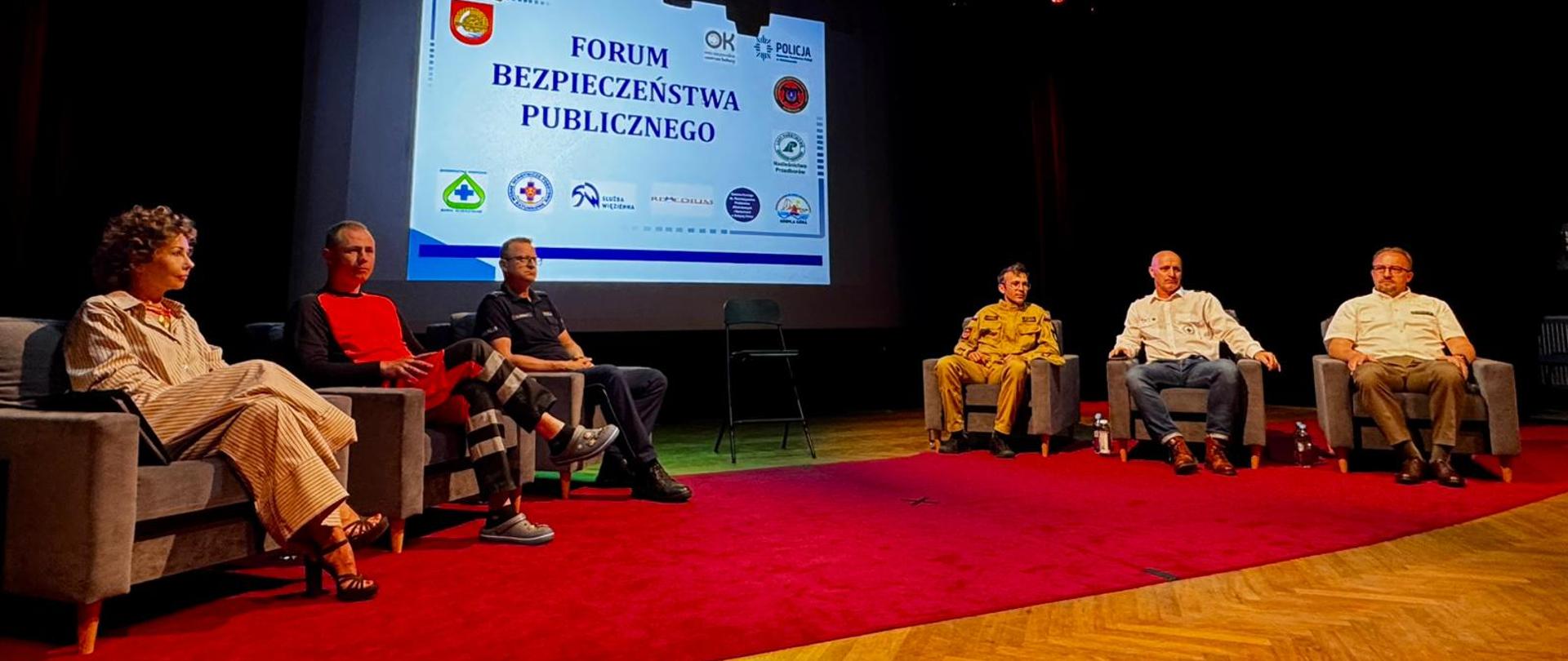 Prelegenci siedzą na scenie w fotelach. Na podłodze czerwony dywan. W tle wyświetlana jest prezentacja