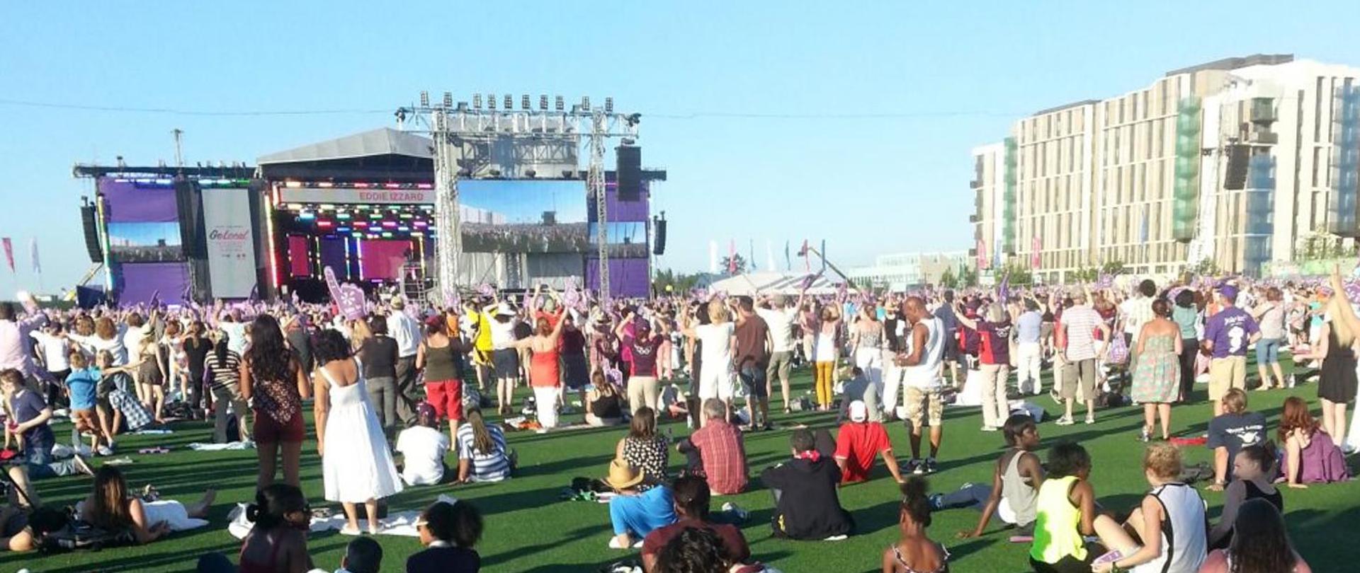 Tłum ludzi uczestniczących w letnim festiwalu plenerowym. W tle scena muzyczna. Po prawej stronie duży blok mieszkalny.