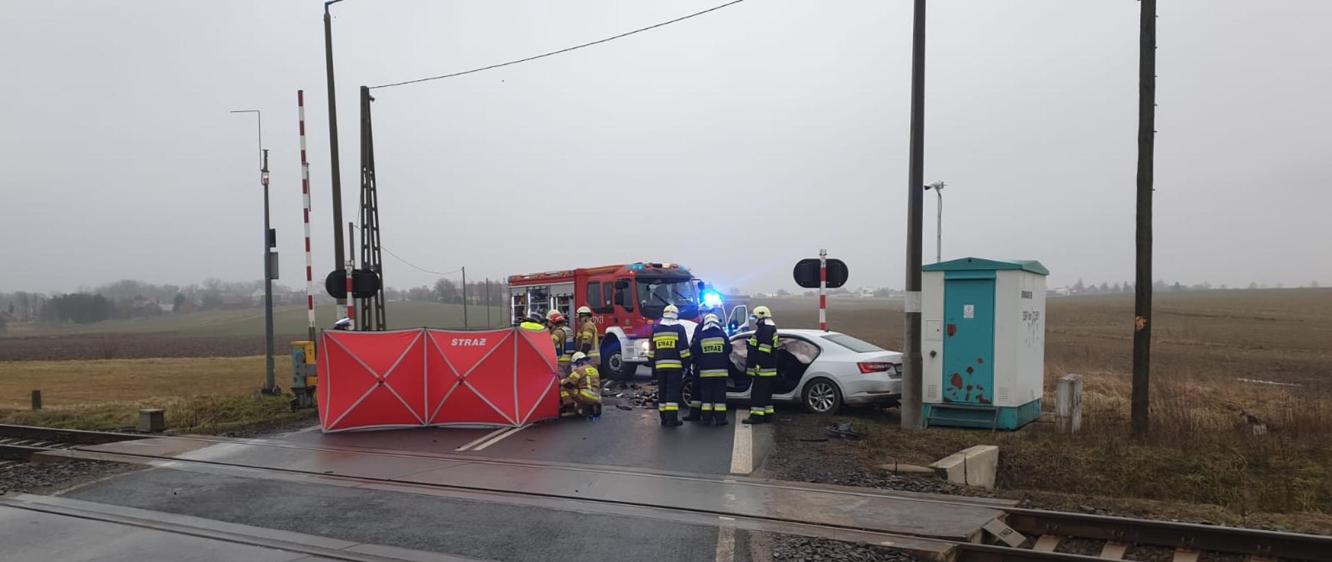 Zdjęcie przedstawia rozbite samochody osobowe po wypadku na przejeździe kolejowym w ciągu drogi krajowej nr 45 w Większycach. Na zdjęciu widoczni strażacy i policjanci pracujący przy zdarzeniu. Jeden z pojazdów zasłonięty parawanem.