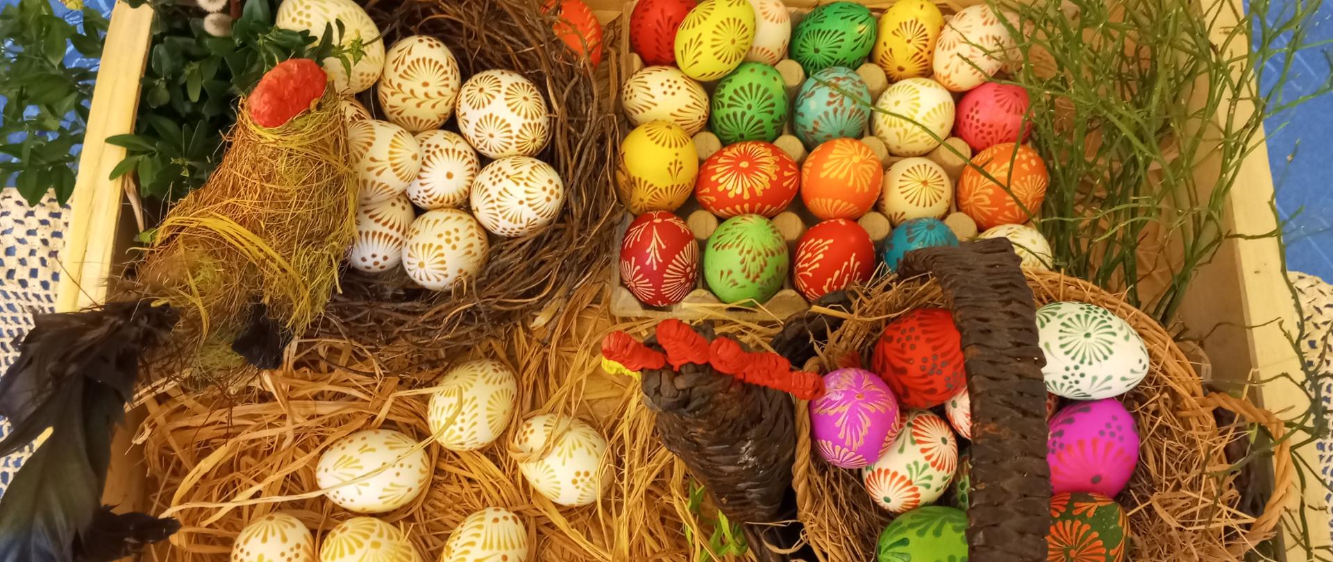 Skrzynia wypełniona kolorowymi pisankami. jest też kogut oraz zielone wiosenne gałązki i bazie
