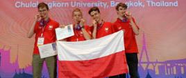 Czterech młodych mężczyzn stoi na scenie, uśmiechając się i prezentując medale oraz flagę Polski. Wszyscy ubrani są w czerwone koszulki polo i mają na szyjach niebieskie wstążki z medalami. Dwóch z nich trzyma dyplomy, a jeden trzyma dyplom oraz flagę Polski, której trzon podtrzymuje mężczyzna po jego lewej. Wszyscy trzymają medale w zębach. W tle widać baner z napisem „26th July – 1st August, 2025” oraz „Chulalongkorn University, Bangkok, Thailand”. Oświetlenie jest jasne, a atmosfera jest radosna i uroczysta.