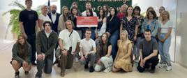 Grupa osób stoi w szkolnym holu i prezentuje tablicę „Szkoła wspierająca honorowe krwiodawstwo”.