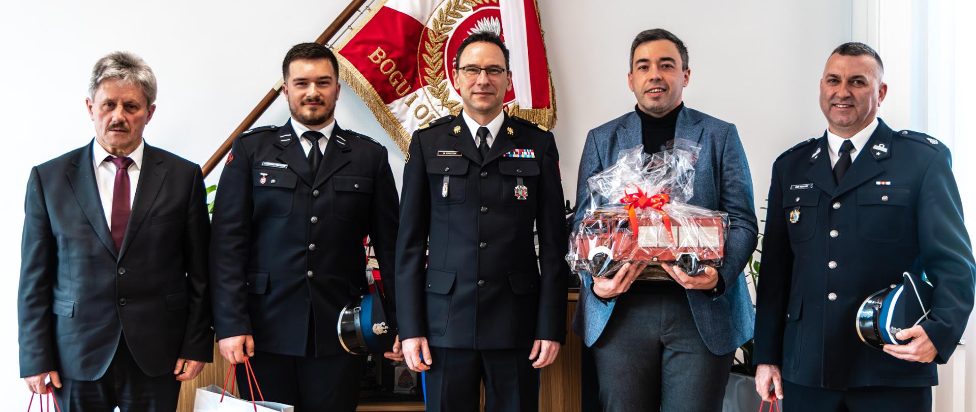 Grupa pięciu mężczyzn, w tym trzech strażaków w mundurach (komendant główny PSP i dwóch strażaków-ochotników) oraz dwóch w stroju cywilnym, pozuje do wspólnego zdjęcia; jedna osoba trzyma model wozu strażackiego, 3 osoby trzymają torby upominkowe.