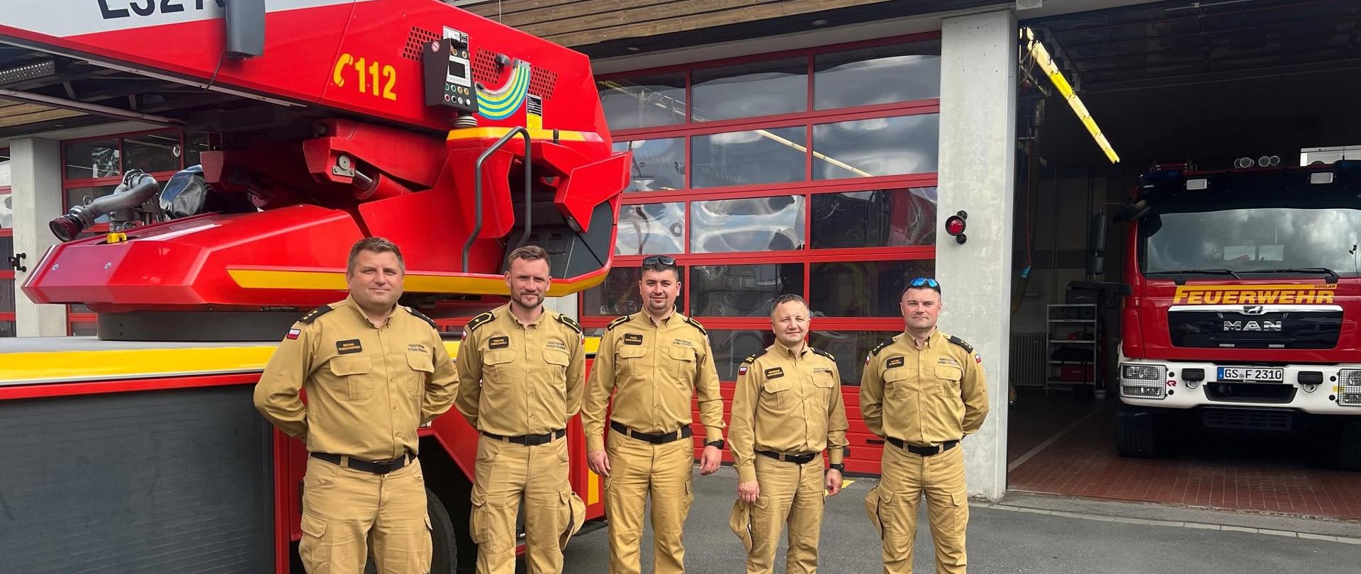 Na zdjęciu przed budynkiem strażackim stoi pięciu strażaków w mundurach w kolorze piaskowym. Mężczyźni są ustawieni w szeregu, patrzą w stronę obiektywu i pozują do fotografii. Po lewej stronie znajduje się czerwony wóz strażacki z wysięgnikiem, na którym widnieje numer alarmowy 112, a w tle, po prawej stronie, w otwartym garażu widoczny jest przód kolejnego czerwonego pojazdu z napisem „FEUERWEHR”.