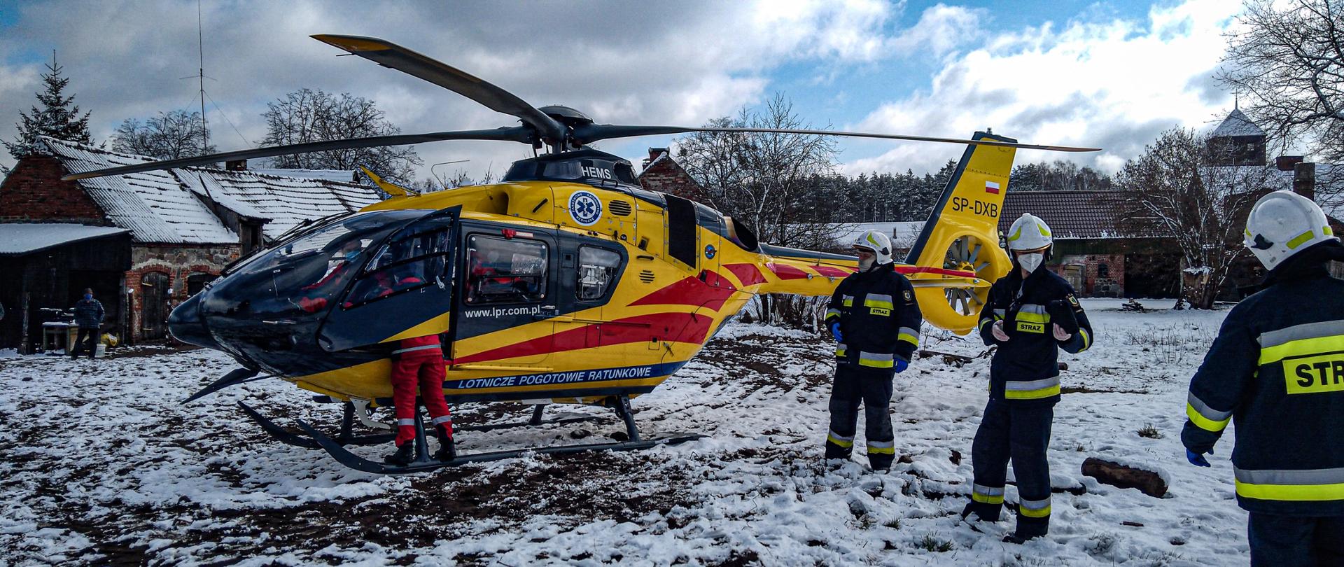 Na zdjęciu znajduje się śmigłowiec Lotniczego Pogotowia Ratunkowego, który jest koloru żółtego. Przy śmigłowcu znajduje się trzech strażaków. Z prawej strony śmigłowca stoi ratownik medyczny, a w środku znajduje się pilot oraz lekarz. Za śmigłowcem są zabudowania gospodarcze. Dookoła leży śnieg. Zdjęcie wykonano w porze dziennej.