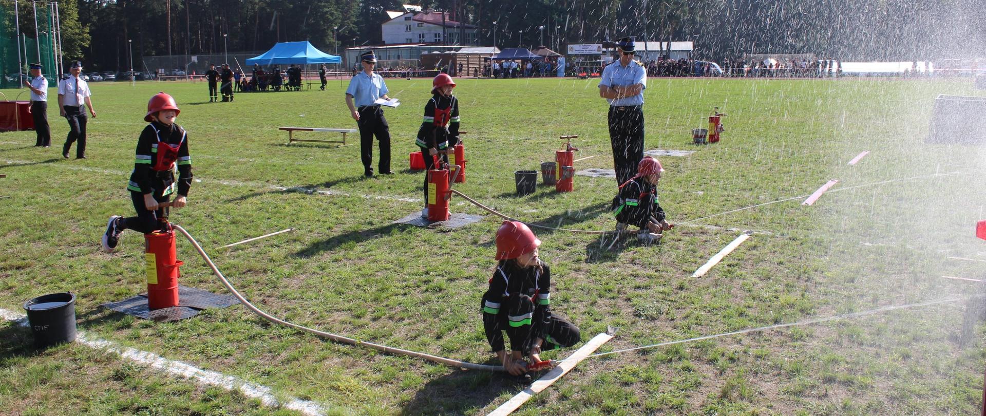 Zawodnicy w ćwiczeniu bojowym MDP, etap napełniania nalewaków z wodą, w tle komisja sędziowska.