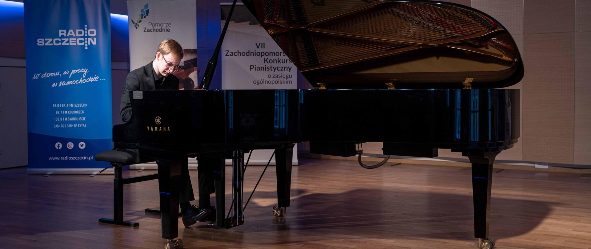 zdjęcie przedstawia wykonawcę grającego na fortepianie na scenie sali koncertowej, w tle banery Radia Szczecin, Pomorza Zachodniego oraz konkursu