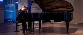 Zdjęcie przedstawia wykonawcę grającego na fortepianie na scenie sali koncertowej, w tle banery Radia Szczecin, Pomorza Zachodniego oraz konkursu