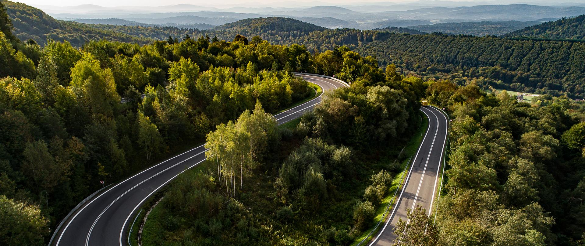 Widok z góry na drogę krajową numer 28, która biegnie w formie serpentyn wśród lasów. Świeci słońce, na drodze jadą auta, a w tle widać zarys gór.