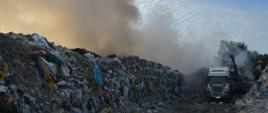 Pożar na składowisku odpadów. Plac składowy. Pomiędzy hałdami odpadów stoi samochód ciężarowy, na który koparka ładuje nadpalone odpady. Panuje silne zadymienie.