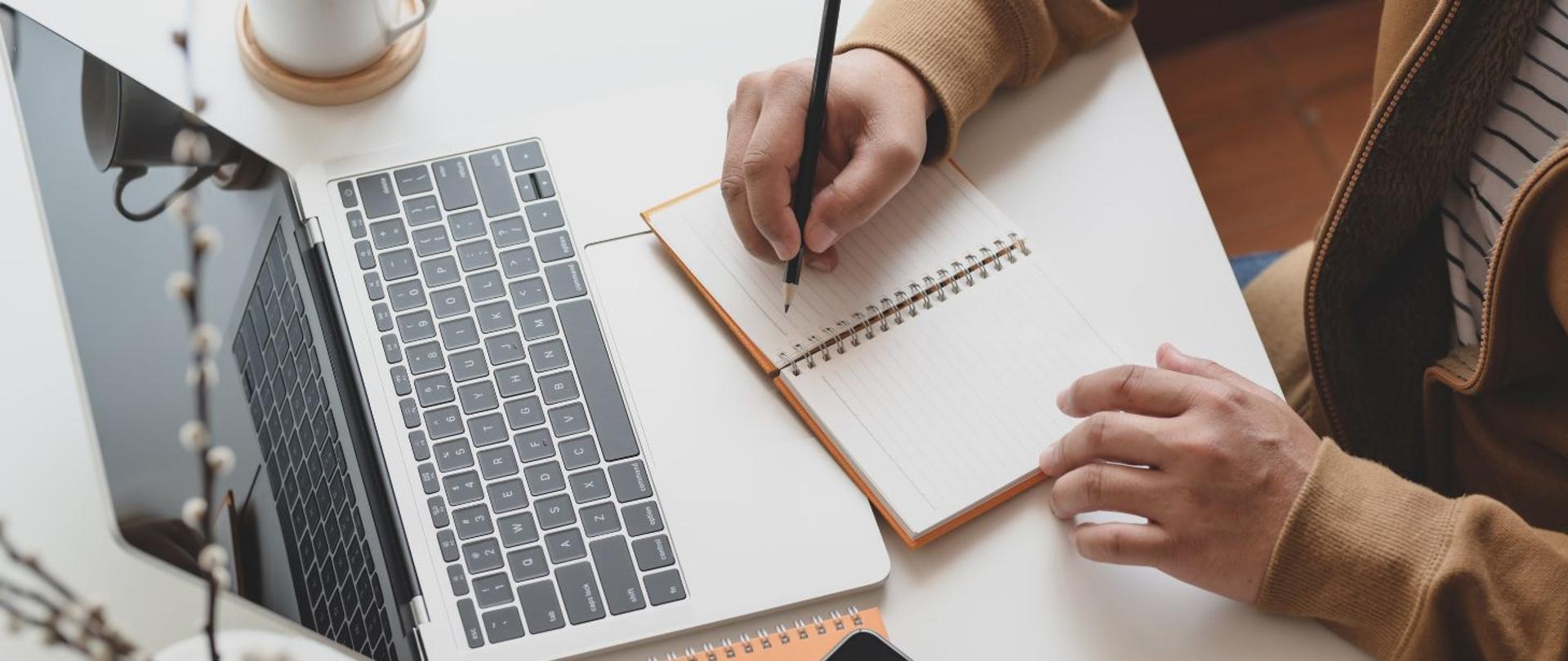 na zdjęciu widoczny jest laptop oraz dłonie osoby, która notuje w notatniku. Na tylnym planie widoczna jest biały kubek. 