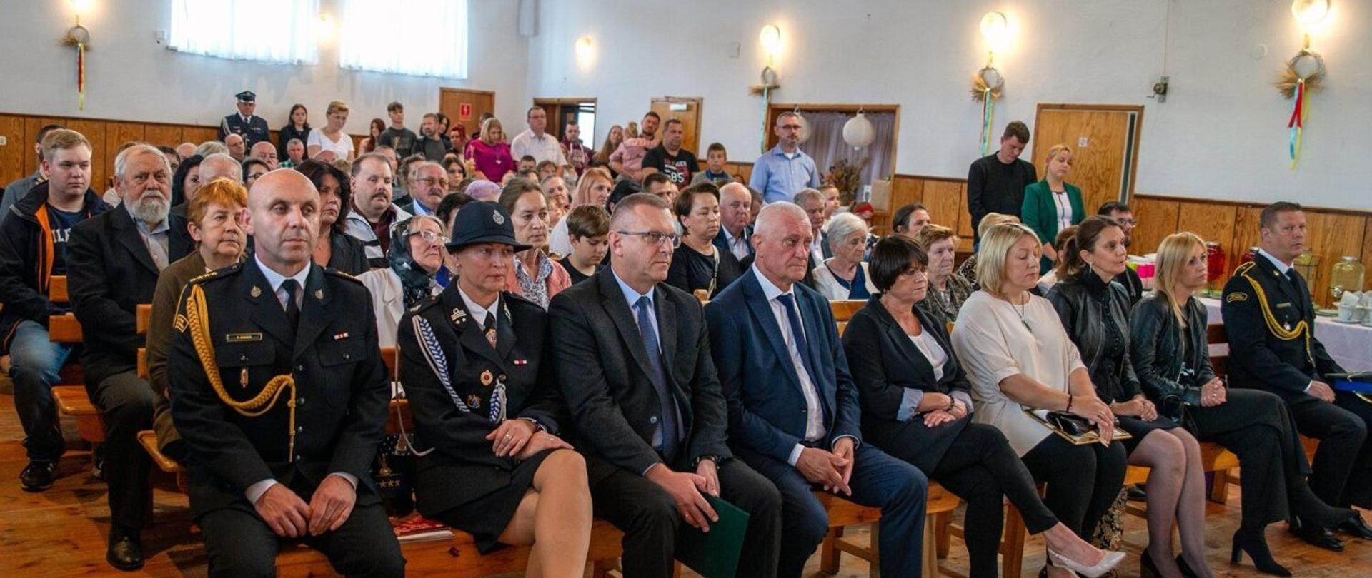 Na oświetlonej sali siedzi grupa mężczyzn i kobiet ubranych w mundury wyjściowe Państwowej i Ochotniczej Straży Pożarnej, oraz osoby w ubraniach cywilnych