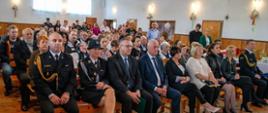 Na oświetlonej sali siedzi grupa mężczyzn i kobiet ubranych w mundury wyjściowe Państwowej i Ochotniczej Straży Pożarnej, oraz osoby w ubraniach cywilnych