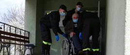 Na zdjęciu strażacy pomagają starszej osobie na wózku inwalidzkim zjechać ze schodów