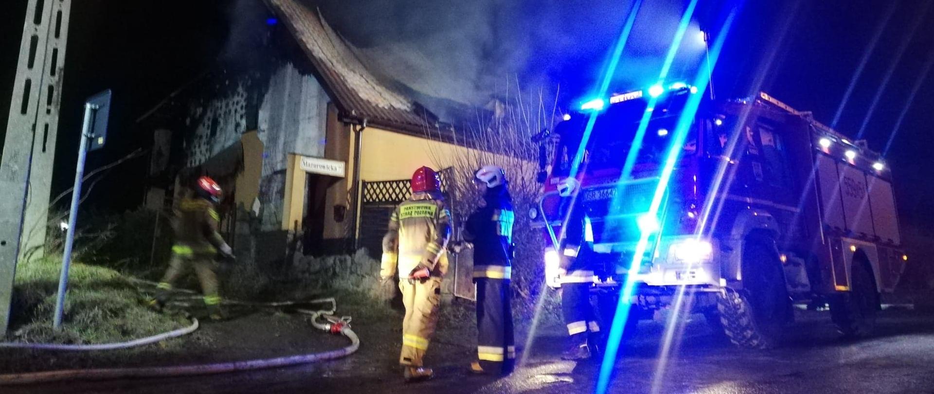 Strażacy stoję przed płonącym budynkiem obok stoi samochód ratowniczo-gaśniczy.
