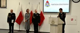 Strażak w mundurze wyjściowym stoi i przemawia przy mównicy za nim stoi kadra kierownicza Państwowej Straży Pożarnej wojewoda pomorska ustawione są flagi Polski na ścianie wisi rota ślubowania ekran z logiem i napisem Państwowej Straży Pożarnej oraz zegar.