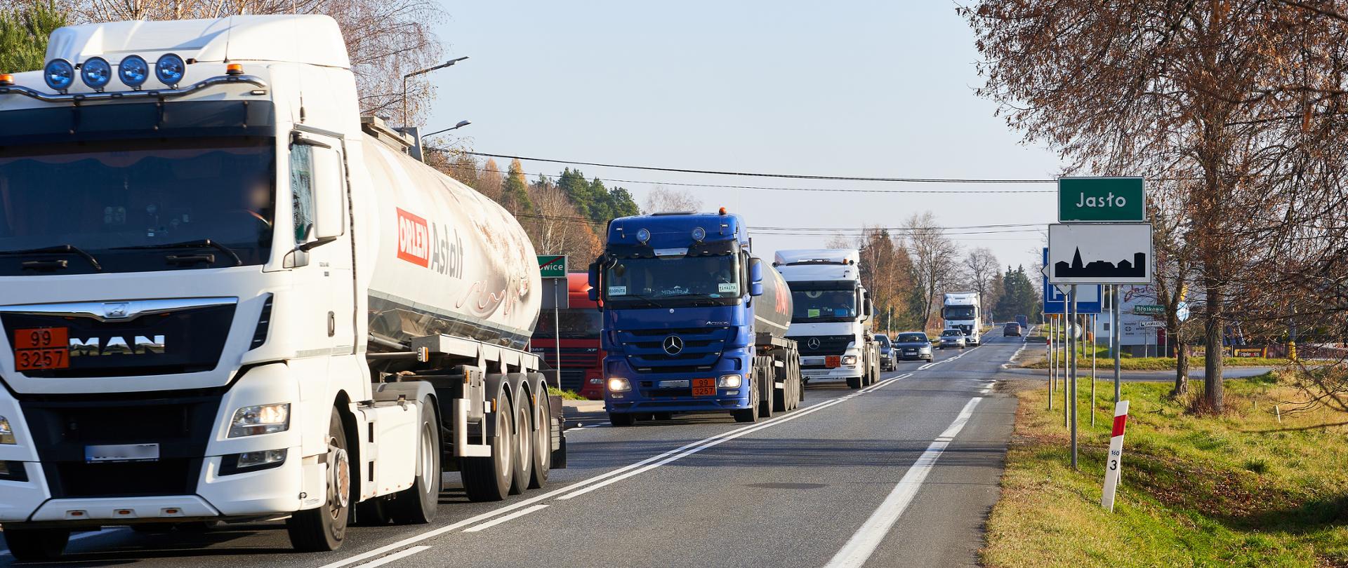 Widok z poziomu drogi na drogę krajową nr 73. Aura wiosenna, świeci słońce. Na jezdni auta ciężarowe i osobowe jadące jedno za drugim, po prawej stronie drogi znak z napisem Jasło. 