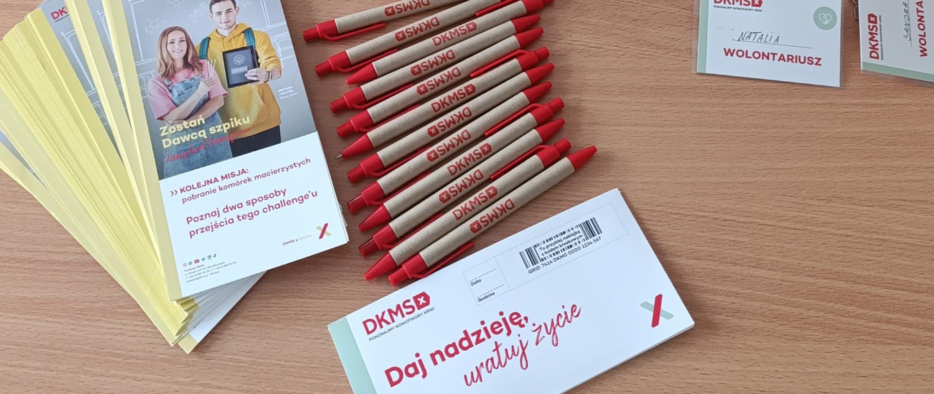 Zestaw materiałów DKMS rozłożonych na stole, w tym smycze, identyfikatory, ulotki, długopisy i pudełko na zgłoszenia.