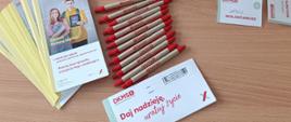 Zestaw materiałów DKMS rozłożonych na stole, w tym smycze, identyfikatory, ulotki, długopisy i pudełko na zgłoszenia.