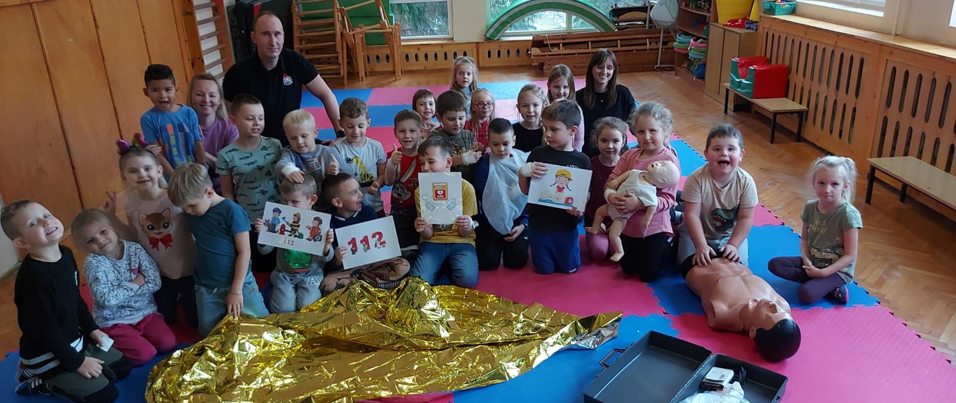 Sala w przedszkolu. Grupka dzieci z panią i strażakiem siedzą na niebiesko różowych matach. Przed nimi leży sprzęt medyczny do pokazów.