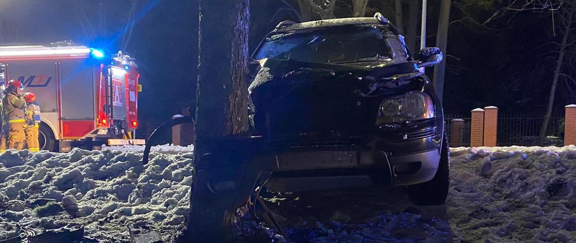 Rozbity samochód osobowy stoi przy drzewie. Ma uszkodzony cały przód. Po lewej stronie stoją strażacy przy samochodzie gaśniczym. Pora nocna. 
