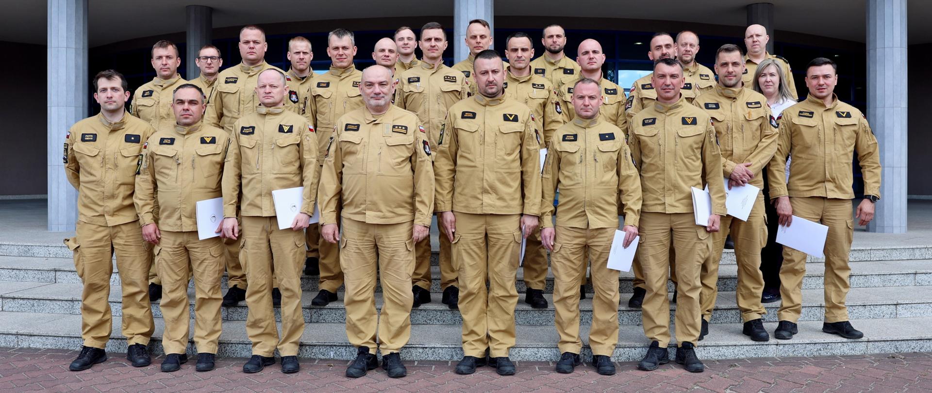 Duża grupa funkcjonariuszy PSP stoi na schodach przed budynkiem, ustawiona do wspólnego zdjęcia. Kilka osób trzyma w rękach dokumenty potwierdzające ukończenie szkolenia. W tle widoczne są kolumny budynku oraz wejście główne. Uczestnicy stoją w kilku rzędach i patrzą w stronę aparatu