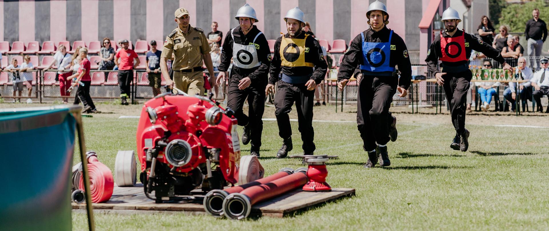 ćwiczenie bojowe rozgrywane na boisku sportowym w Brzezinach, do motopompy na podeście dobiegają zawodnicy OSP Polik i będą rozwijać odcinki w celu strącenia wodą pachołków 
