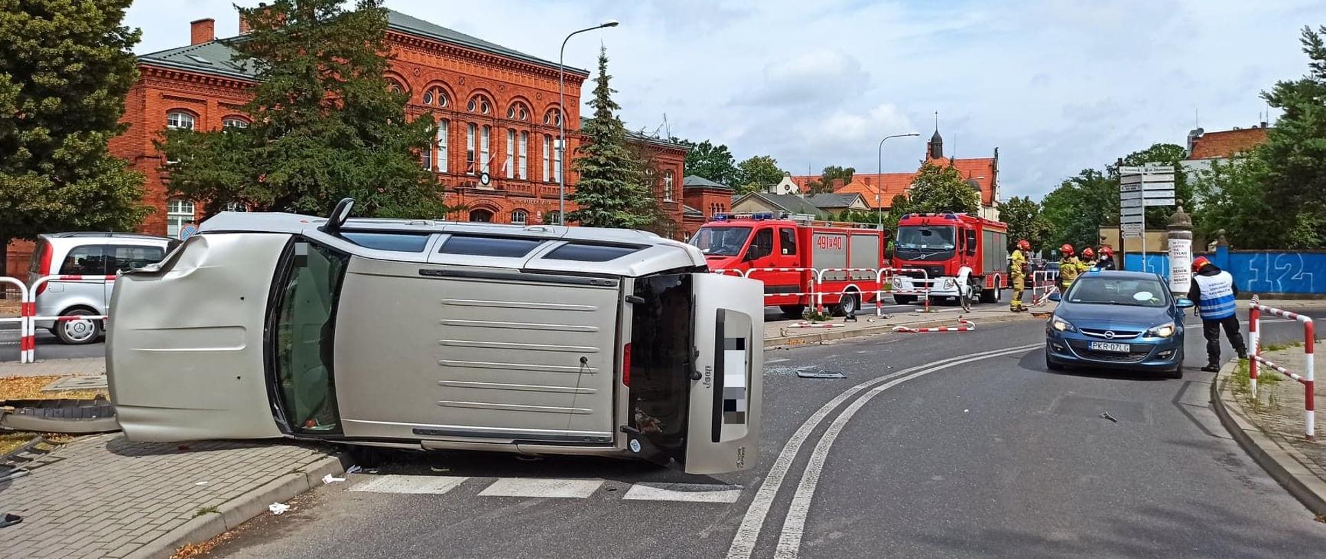 W wyniku wypadku samochód osobowy przewrócił się na bok i blokuje jeden pas drogi krajowej. Strażacy podejmują czynności na miejscu zdarzenia oczyszczając nawierzchnię drogi z elementów karoserii samochodowej. Na drugim pasie jezdni zaparkowany jest radiowóz policyjny. Dwa wozy straży pożarnej zaparkowane zostały po drugiej stronie chodnika. 