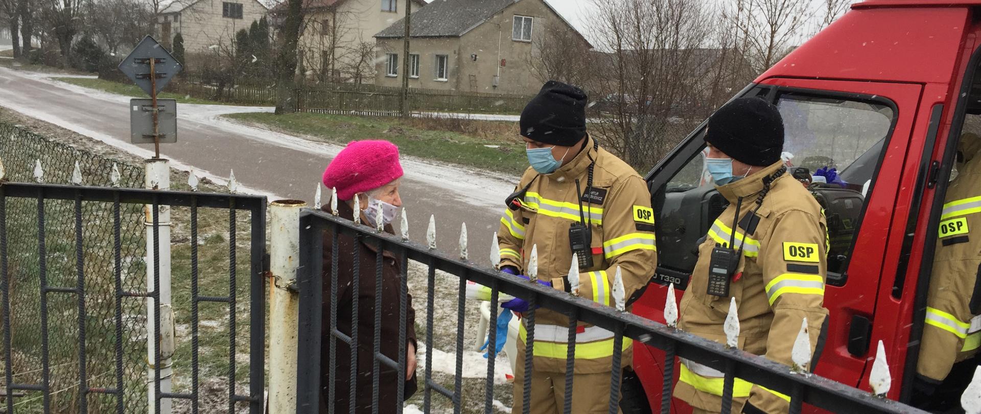Dwóch strażaków Ochotniej Straży Pożarnej stoją w żółtych ubraniach specjalnych w maseczkach i rękawiczkach lateksowych na chodniku przed bramą posesji ze starszą kobietą ubraną na czarno w różowym berecie. Przy strażakach stoi czerwony samochód OSP. W tle domy mieszkalne i drzewa.