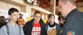Targi edukacyjne. Sala sportowa w ZSZ w Rawiczu. Strażak prezentuje uczniom zasady naboru do służby w PSP oraz zasady rekrutacji do szkół pożarniczych. Trzyma w rękach informatory wydane przez Szkołę Główną Służby Pożarniczej. 