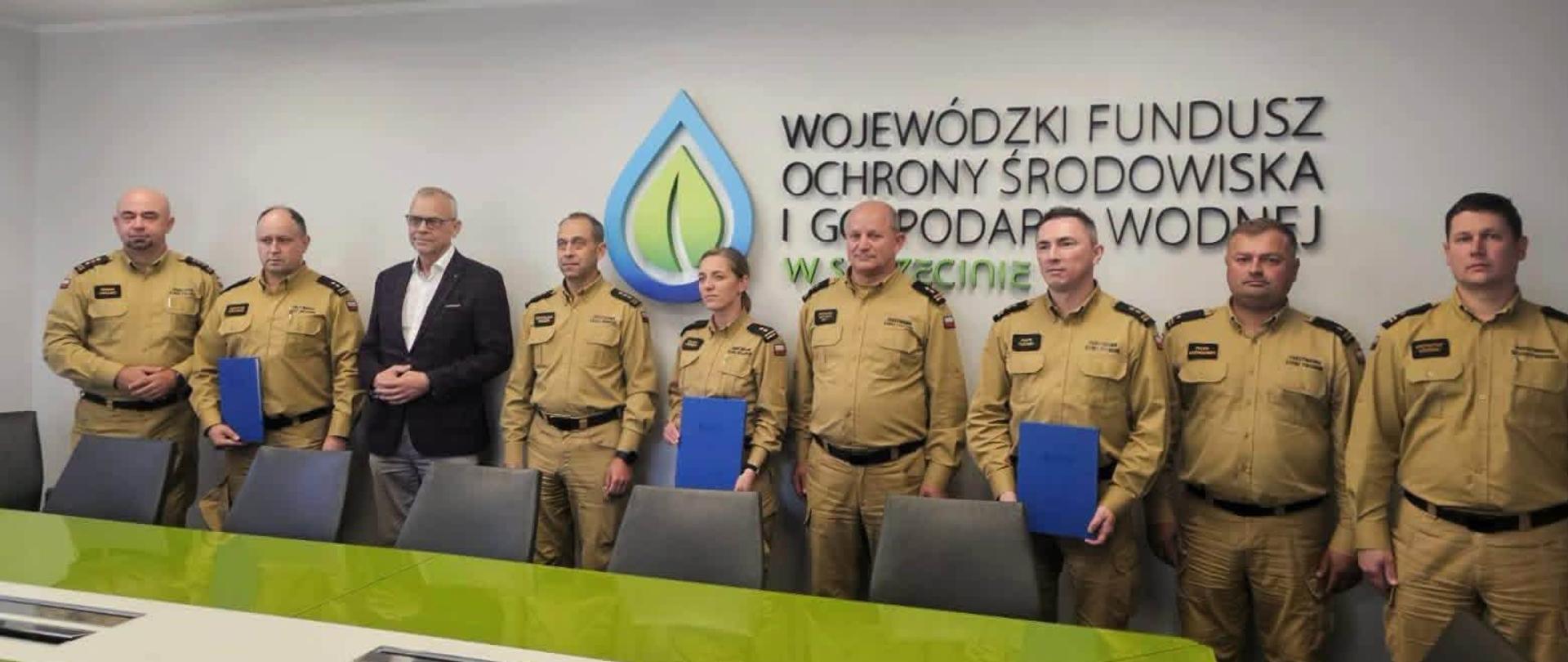 Grupa funkcjonariuszy Państwowej Straży Pożarnej w piaskowych mundurach galowych stoi w sali konferencyjnej Wojewódzkiego Funduszu Ochrony Środowiska i Gospodarki Wodnej w Szczecinie. Wśród uczestników znajduje się mężczyzna w garniturze. Kilka osób trzyma niebieskie teczki. W tle widoczne jest logo i nazwa instytucji.
