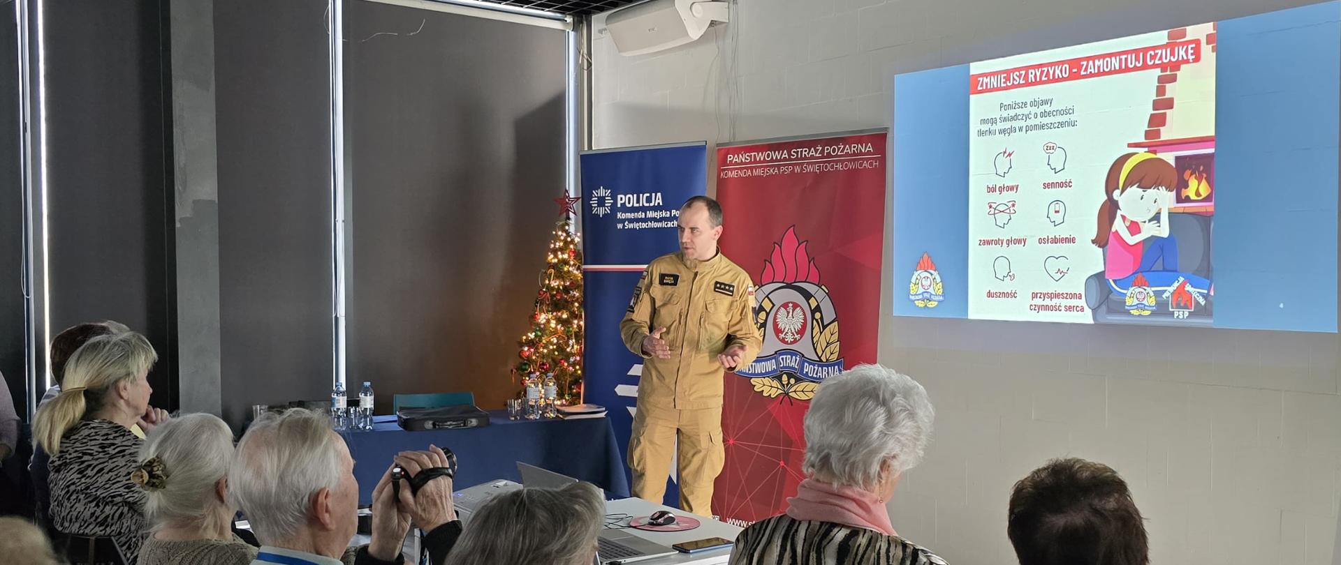 Zdjęcie przedstawia spotkanie edukacyjne w nowoczesnej sali, podczas którego strażak w piaskowym mundurze prowadzi prelekcję dla grupy osób. Funkcjonariusz stoi na tle banerów Policji oraz Państwowej Straży Pożarnej, omawiając zagadnienia bezpieczeństwa. Obecność choinki w tle sugeruje okres zimowy, co bezpośrednio wiąże się z tematyką spotkania – zwiększonym ryzykiem pożarów i zatruć w sezonie grzewczym.
Kluczowym elementem jest wyświetlana na ścianie prezentacja zatytułowana „ZMNIEJSZ RYZYKO – ZAMONTUJ CZUJKĘ”. Slajd w przejrzysty sposób obrazuje objawy zatrucia tlenkiem węgla, takie jak ból głowy, duszności czy senność. Słuchacze siedzą na białych krzesłach, z uwagą śledząc wystąpienie i wyświetlane grafiki, które mają na celu podniesienie ich świadomości na temat domowych zagrożeń.
