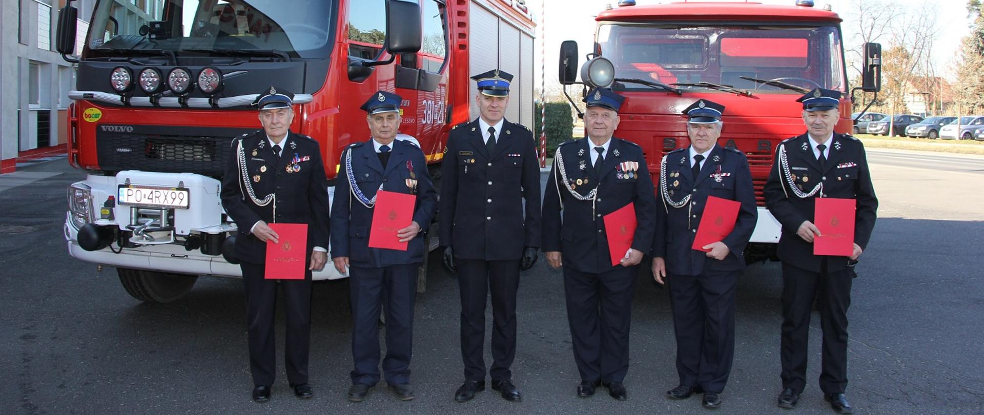 Osoby w mundurach stoją w szeregu. W tle widoczne pojazdy pożarnicze oraz budynek komendy.
