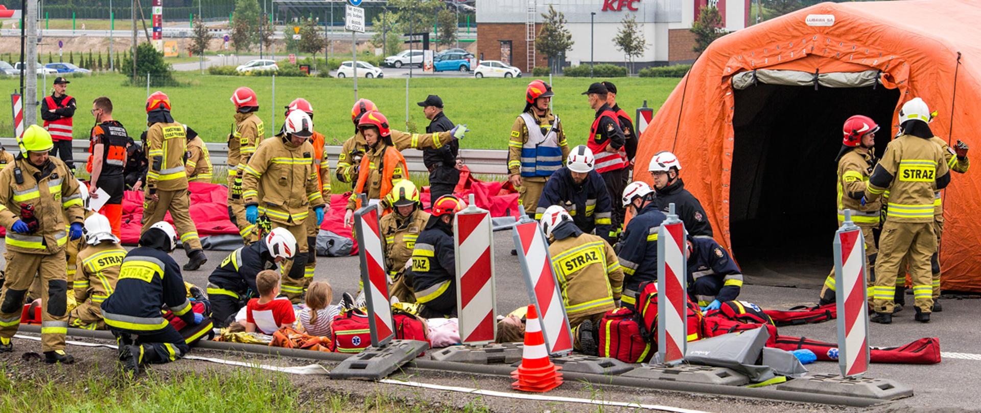 Na budowanej nitce drogi S5 po prawej widać stojący namiot pneumatyczny, wokół niego znajduje się kilkudziesięciu strażaków ubranych w ubrania specjalne oraz hełmy, na ziemi znajdują się poszkodowani, przy których cześć strażaków klęczy i udziela pomocy przedlekarskiej.
