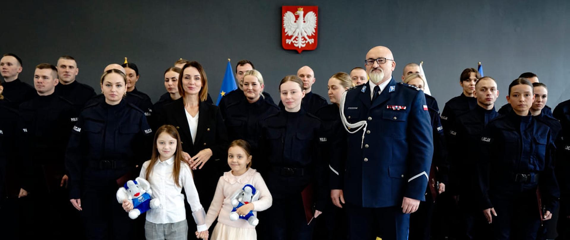 grupa policjantów stoi na ścianie, na której wisi godło, jest tez kobieta (wojewoda) i dwoje dzieci