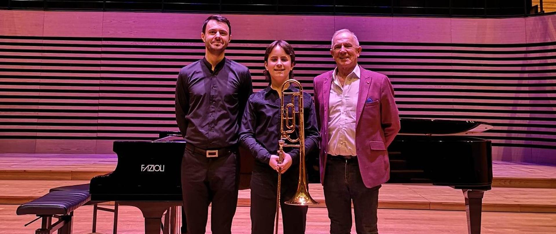 Na scenie koncertowej na tle czarnego fortepianu stoją: nauczyciel akompaniator, uczeń z puzonem oraz nauczyciel instrumentu. W tle znajdują sie rzędy foteli dla publiczności. U góry zawieszony ekran z nazwą koncertu.