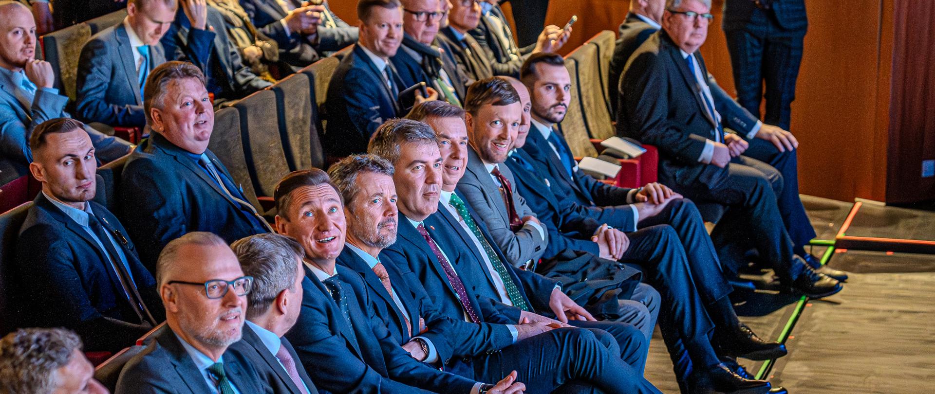 Widok z boku na salę, w rzędach krzeseł siedzą ludzie, w pierwszym rzędzie minister wieczorek, król Danii i kilku mężczyzn w garniturach.