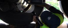 Inspektor transportu Drogowego leży pod pojazdem, między dwoma kołami z tej samej strony pojazdu. Przy pomocy zapalonej latarki sprawdza stan zużycia tarcz hamulcowych