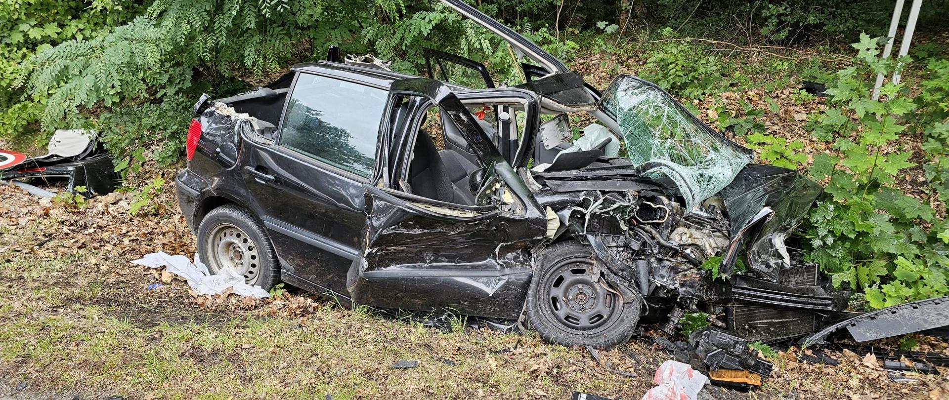 Czarny samochód osobowy marki VW Polo po zderzeniu z innym pojazdem, znajduje się na poboczu drogi w Łęknicy. dookoła porozrzucane elementy pojazdu.