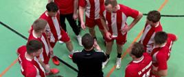 Wnętrze hali sportowej SA PSP w Krakowie. Zdjęcie zrobione z góry – w na parkiecie ustawieni w kręgu naradzający się przed meczem zawodnicy - mężczyźni w strojach sportowych – większość w biało-czerwonych barwach.