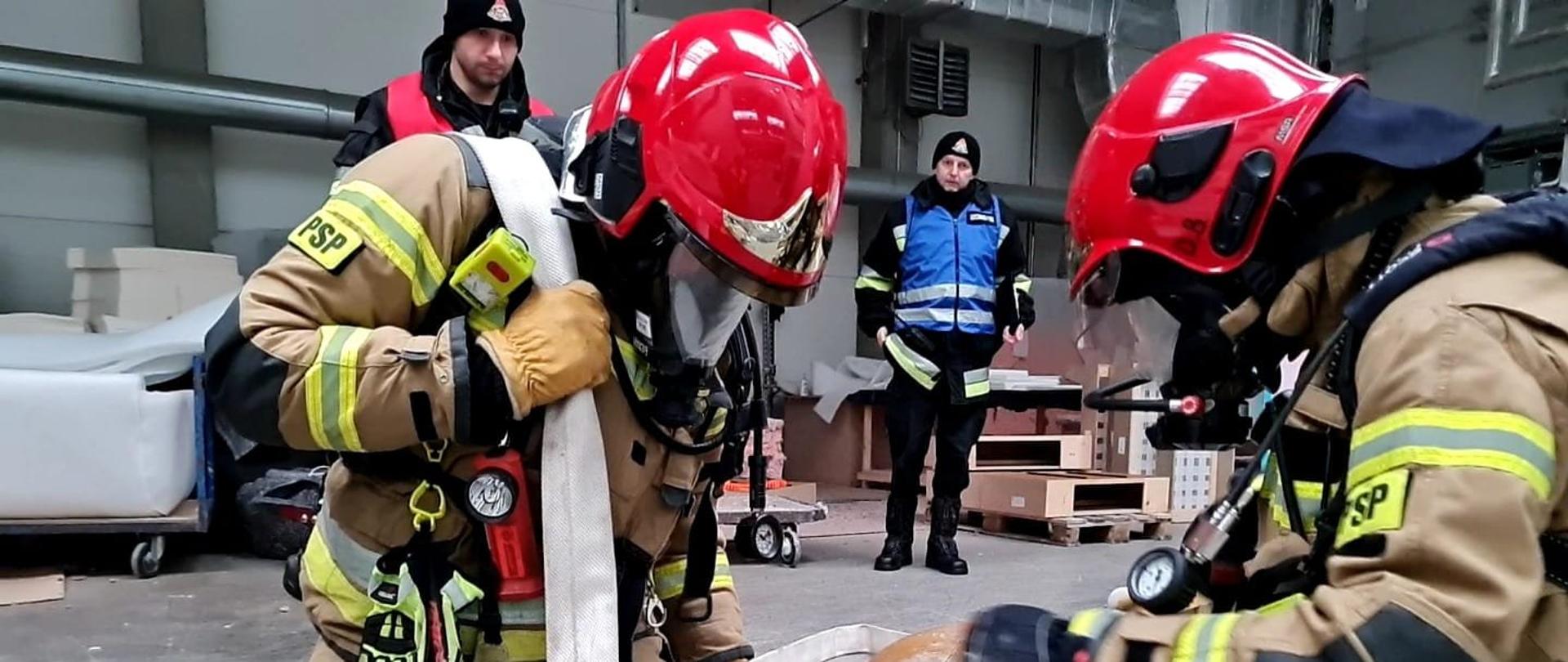 Strażacy ewakuują manekina, poruszając się na kolanach z przewieszoną linią wężową przez ramię. Strażacy ubrani w piaskowe ubranie specjalne i czerwony hełm.