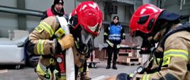 Strażacy ewakuują manekina, poruszając się na kolanach z przewieszoną linią wężową przez ramię. Strażacy ubrani w piaskowe ubranie specjalne i czerwony hełm 