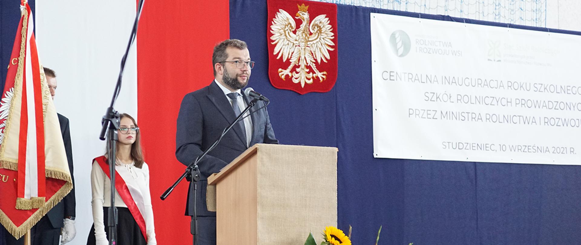 Minister Grzegorz Puda stojący przy mównicy podczas rozpoczęcia roku szkolnego, obok poczet sztandarowy ze sztandarem szkoły.