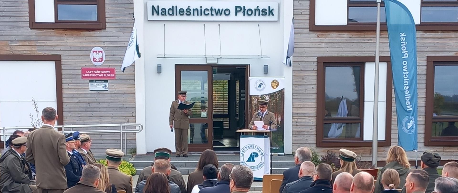 Na zdjęciu widać uroczystość plenerową przed budynkiem Nadleśnictwa Płońsk. Przed wejściem stoi umundurowany leśnik, przy mównicy z logo Lasy Państwowe przemawia drugi. Na krzesłach siedzi kilkadziesiąt osób, głównie w mundurach leśnych. W tle widoczne są flagi i tablice z nazwą nadleśnictwa.