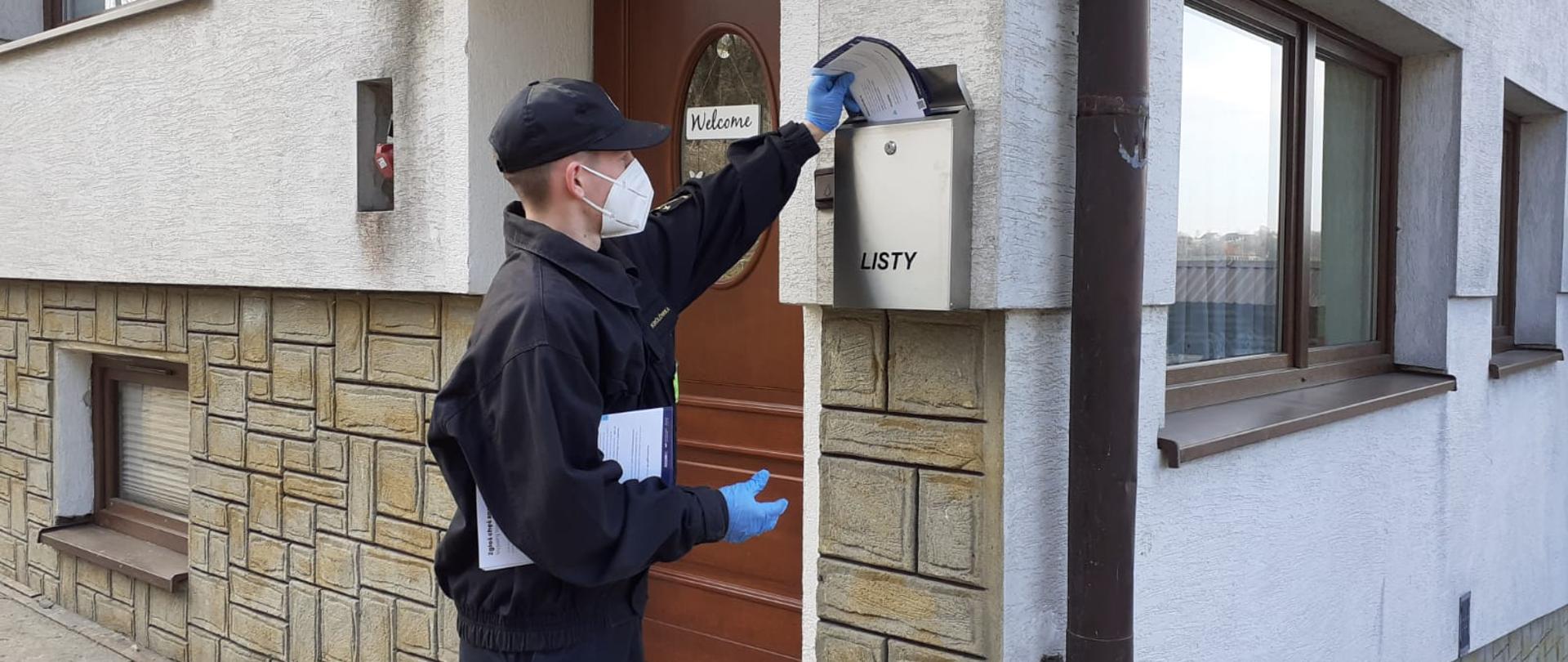 Mężczyzna ubrany w czarny mundur strażacki z odblaskowymi paskami, czarna czapką na głowie, czarnym butami, na rękach ma założone rękawiczki lateksowe koloru niebieskiego, na twarzy ma ubrana maseczkę chirurgiczną koloru białego, stoi on przed wejściem do budynku. Na ścianie przy wejściu znajduje się skrzynka na listy w kolorze srebrnym. W rękach trzyma ulotkę informacyjną która wkłada do skrzynki na listy. Budynek w kolorze białym, okna i drzwi koloru brązowego, wejście do budynku po schodach betonowych.