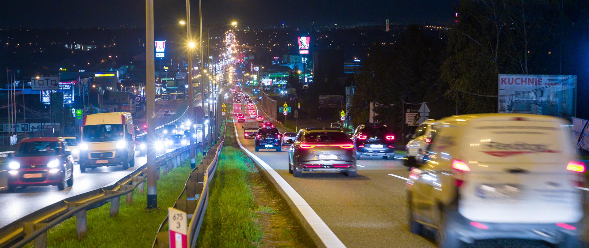 Zdjęcie przedstawia nocny widok na drogę krajową DK7 w Libertowie pod Krakowem. Widać na nim dwupasmową jezdnię z dużym natężeniem ruchu, zarówno w kierunku Krakowa, jak i od Krakowa. Droga jest oświetlona latarniami ulicznymi, a w tle można dostrzec liczne reklamy świetlne oraz budynki. Na pierwszym planie, po lewej stronie, znajduje się barierka ochronna.