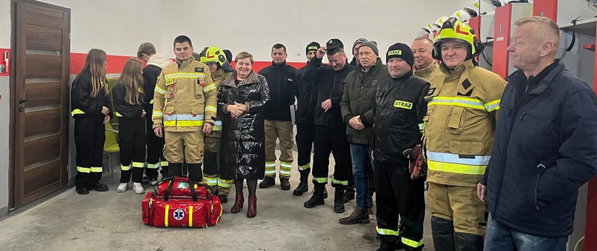 W remizie strażackiej Dyrektor OR KRUS stoi obok Prezesa OSP, przed nimi leży torba z zestawem ratownictwa z tyłu stoją strażacy