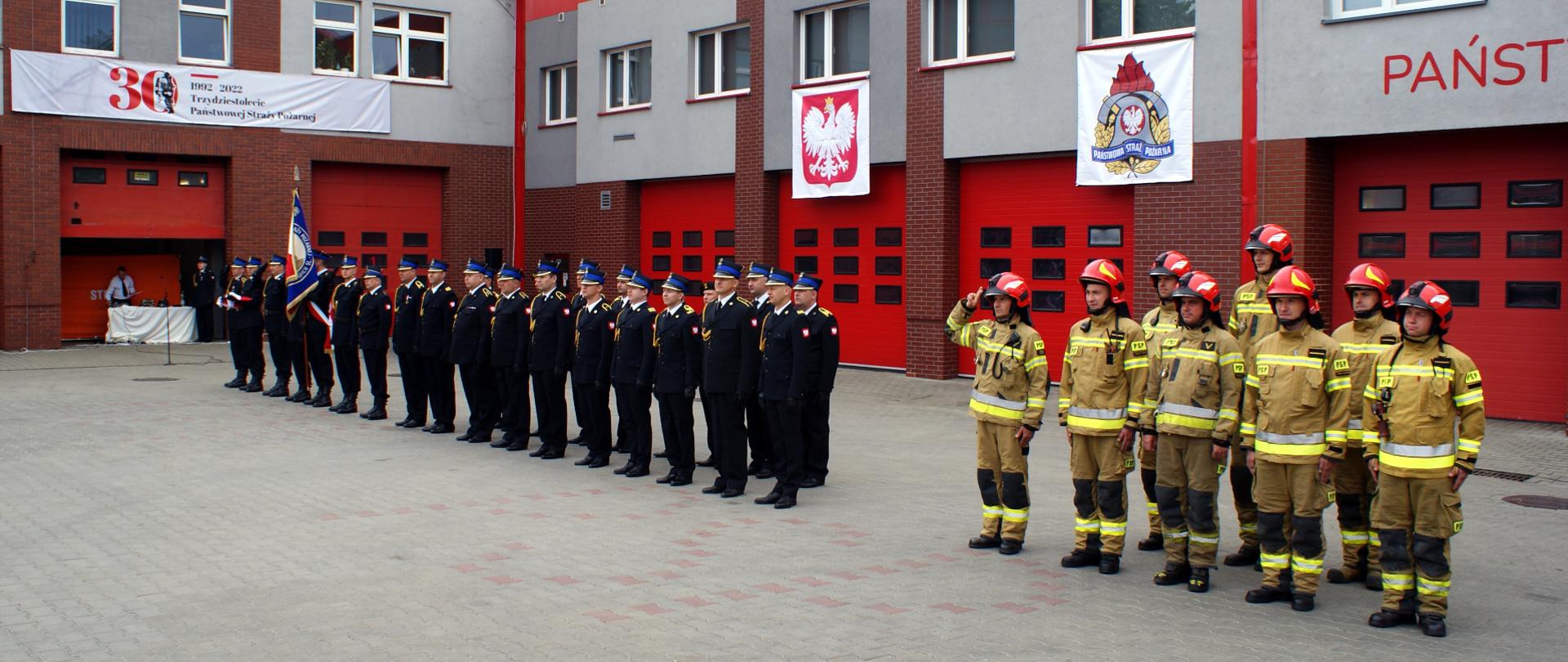 Zdjęcie przedstawia dwa pododdziały strażaków. Jeden w mundurach wyjściowych a drugi w ubraniach specjalnych. Obok stoi poczet flagowy i sztandarowy. W tle budynek Komendy Miejskiej PSP w Piekarach Śląskich.