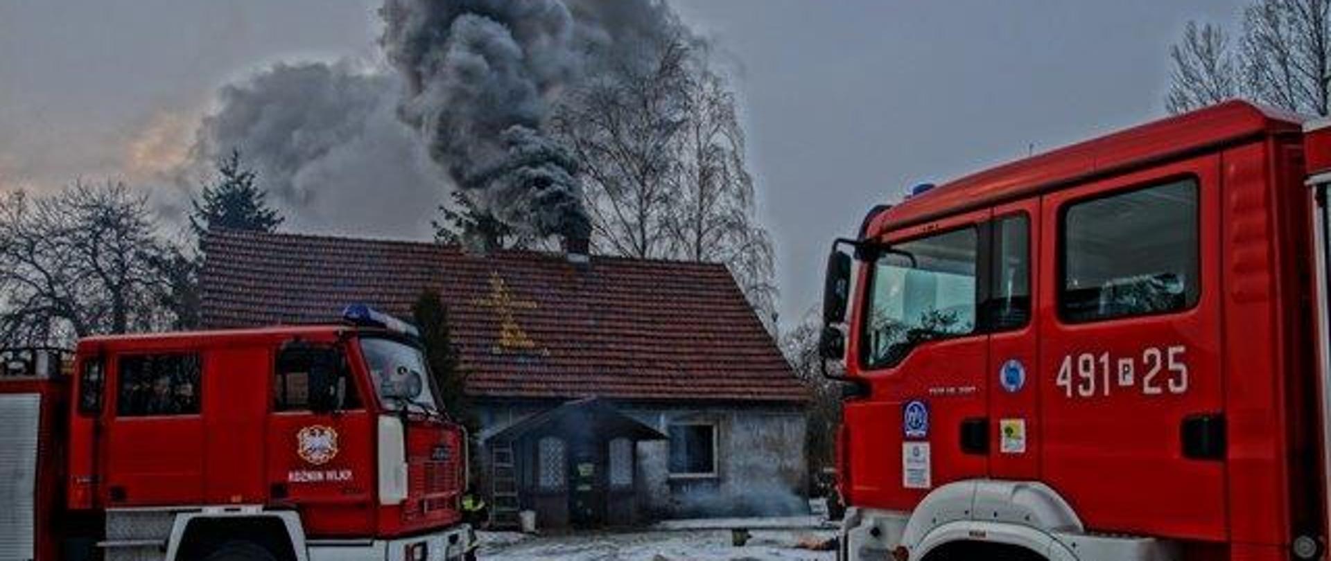 Z komina budynku jednorodzinnego wydobywa się gęsty dym. Zimowa sceneria. Ziemia pokryta śniegiem. Dach budynku spadzisty pokryty dachówką. Przed budynkiem stoją dwa wozy pożarnicze .