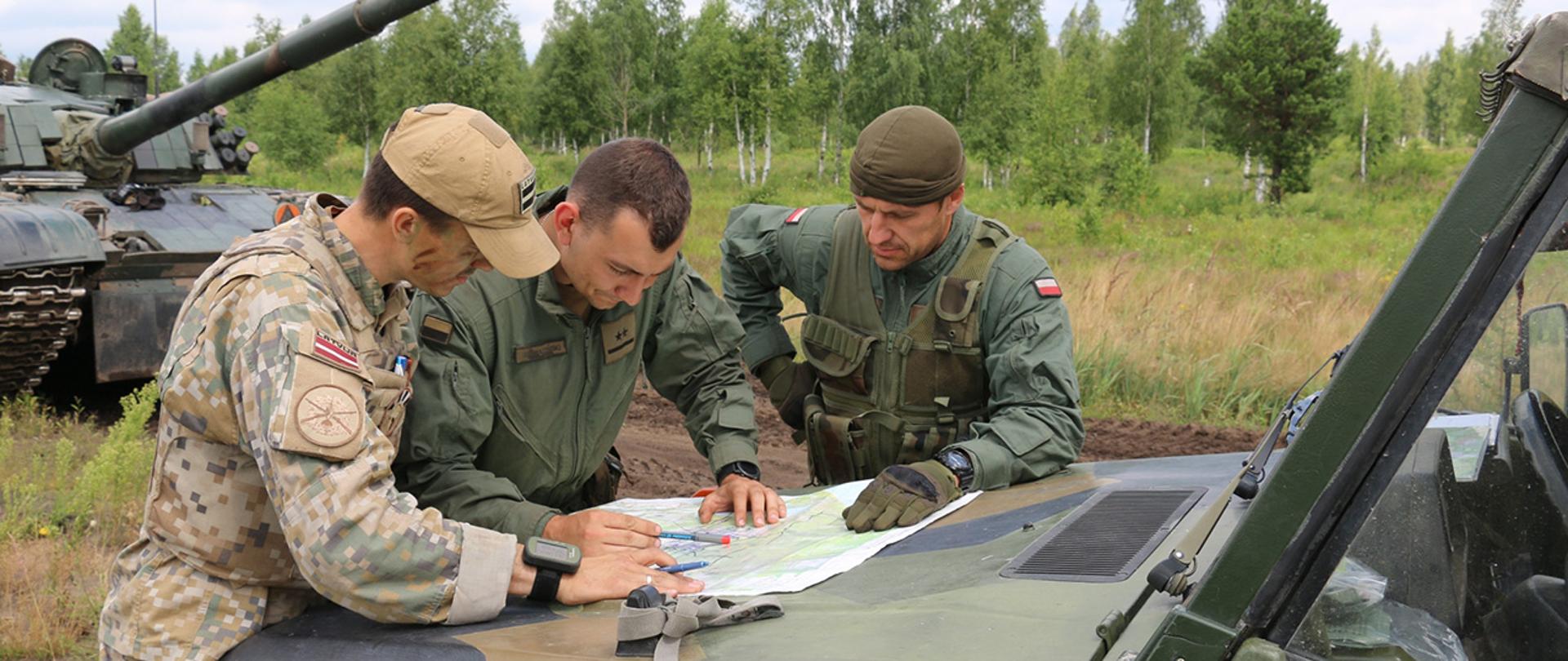Żołnierze Łotewscy i Polscy sprawdzają swoje położenie na mapie. W tle zieleń oraz sprzęt wojskowy.